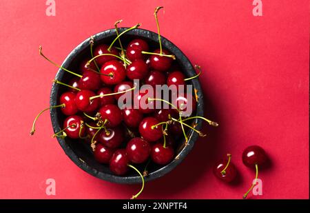 Une image frappante représentant un bol noir rempli de cerises fraîches et mûres sur un fond rouge foncé. Le contraste vif entre le bol sombre Banque D'Images