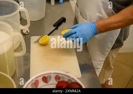 Une personne portant des gants bleus coupe un citron sur une planche à découper dans une cuisine. La scène comprend un bol de fraises et divers ustensiles de cuisine Banque D'Images