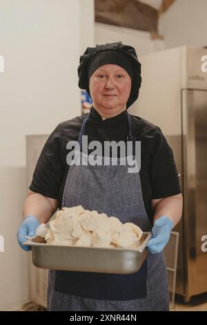 Une femme chef portant un chapeau de chef noir et des gants bleus, tenant un plateau rempli de pain fraîchement cuit. Elle se tient dans une cuisine en acier inoxydable Banque D'Images