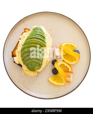 Gros plan de pain grillé à l'avocat garni d'œufs brouillés, servi avec un accompagnement de fruits frais comprenant des oranges, des fraises et des bleuets. Banque D'Images