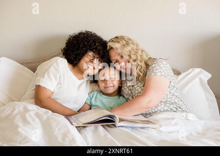 Mère et filles câlinent dans le lit avec livre sur les genoux Banque D'Images