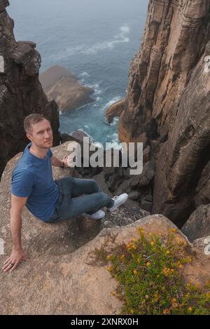 Homme assis sur Cliff Edge accidenté surplombant Misty Ocean au Portugal Banque D'Images