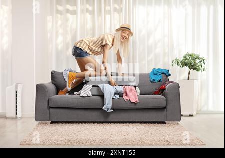 Jeune femme joyeuse essayant de fermer une valise pleine de vêtements sur le dessus d'un canapé à la maison Banque D'Images