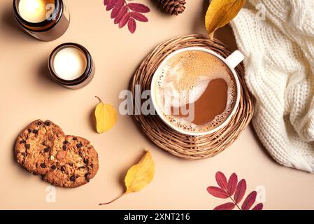 Concept hygge d'automne. Tasse de café cappuccino, biscuits aux pépites de chocolat, bougies allumées et feuilles jaunes sur fond beige, vue de dessus. Confort douillet Banque D'Images
