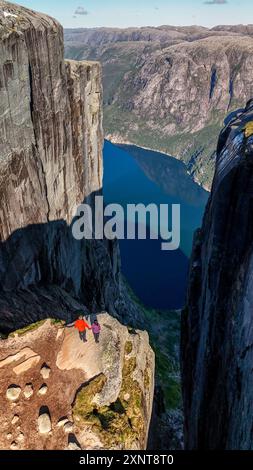 Deux randonneurs se tiennent au bord d'une falaise spectaculaire, contemplant le fjord bleu profond en contrebas et les formations rocheuses imposantes autour d'eux sous le ciel lumineux. Kjerag Kjeragbolten Lysefjord Norvège Banque D'Images