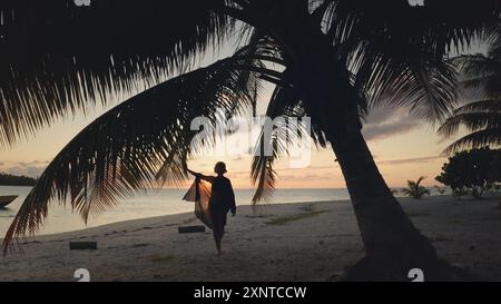 Femme gracieusement debout sur une plage magnifique aux maldives, se prélasser dans la lueur chaude du coucher de soleil, entourée de palmiers se balançant doucement dans la brise tropicale Banque D'Images