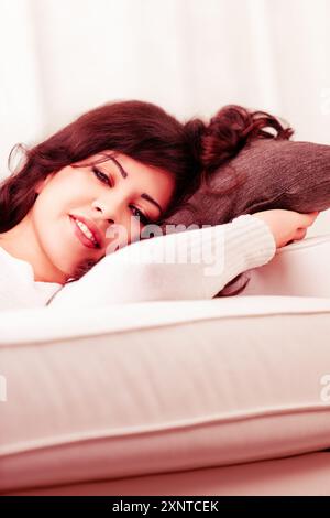 Jeune femme se détend sur un canapé confortable à la maison, à l'air paisible et serein dans un salon élégant, rêvant et souriant joyeusement Banque D'Images