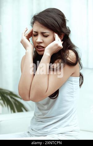 Jeune femme assise sur un canapé à la maison, tenant ses tempes dans la douleur, montrant le stress et la tension. Elle a l'air malheureuse et inquiète, reflétant l'effet migraineux Banque D'Images