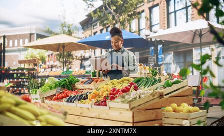 Portrait d'une vendeuse de rue africaine multiethnique travaillant sur un ordinateur portable tout en se tenant debout dans un stand de fermiers avec des produits agricoles biologiques frais. Femme d'affaires contactant les fournisseurs en ligne Banque D'Images