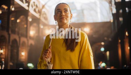 Jeune femme en chandail jaune contemplant dans une bibliothèque historique, tenant un livre. Étudiante intelligente explorant l'ambiance académique, entourée d'architecture classique et d'éclairage doux Banque D'Images