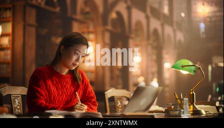 Etudiante asiatique engagée dans l'étude avec des livres et ordinateur portable dans le cadre de bibliothèque classique. Jeune femme concentrée sur le E-Learning, entourée d'architecture classique et de meubles en bois riche. Banque D'Images