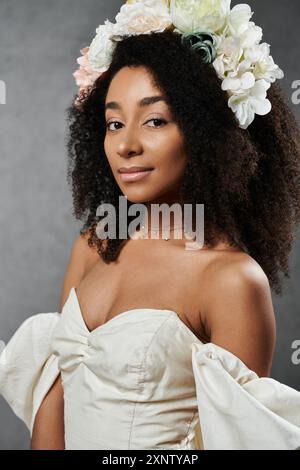 Une belle mariée afro-américaine dans une robe de mariée blanche avec des fleurs dans ses cheveux pose sur un fond gris. Banque D'Images