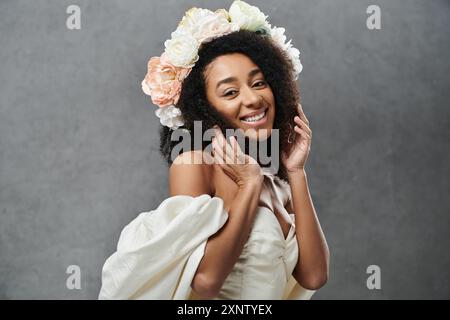 Une belle mariée afro-américaine dans une robe de mariée blanche sourit radieusement, portant une couronne florale dans ses cheveux sur un fond gris. Banque D'Images