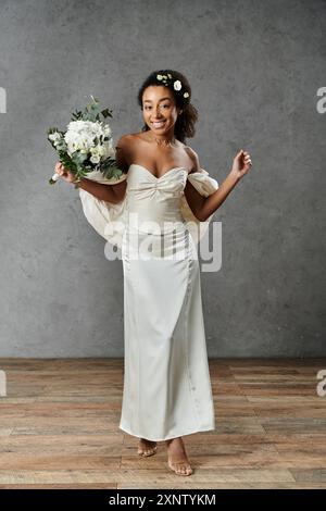 Une belle mariée afro-américaine dans une robe de mariée blanche avec des fleurs dans ses cheveux, debout sur un fond gris. Banque D'Images