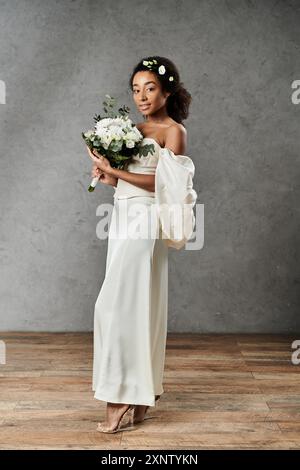 Une belle mariée afro-américaine dans une robe de mariée blanche tient un bouquet de fleurs et sourit à la caméra. Banque D'Images