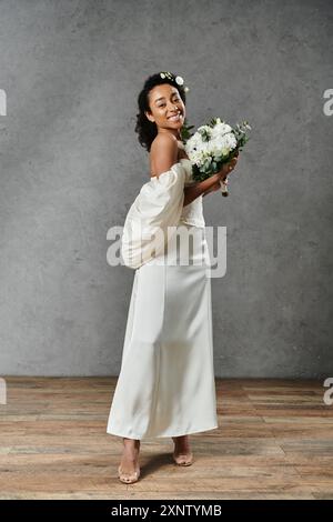 Une belle mariée afro-américaine dans une robe de mariée blanche avec des fleurs dans ses cheveux, souriant et tenant un bouquet. Banque D'Images