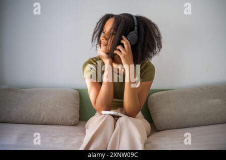 Femme contemplative afro-américaine écoutant de la musique avec des écouteurs, assise sur le canapé. Banque D'Images