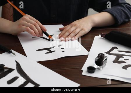 Calligraphie. Femme avec pinceau et encrier pratiquant l'écriture de hiéroglyphes chinois sur papier à la table en bois, gros plan Banque D'Images