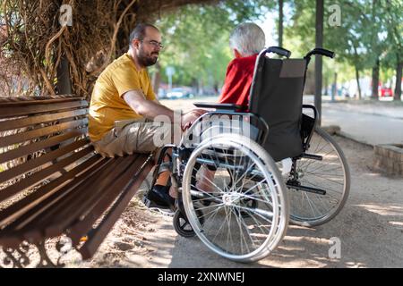 Jeune homme assis sur un banc tout en parlant joyeusement à sa grand-mère aux cheveux blancs assise dans un fauteuil roulant. Banque D'Images