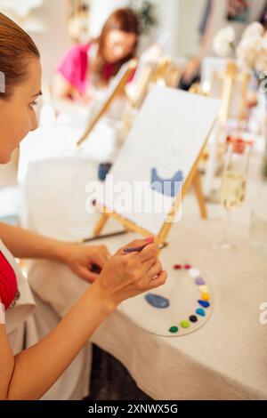 Une main féminine avec un pinceau et une palette avec des peintures. L’artiste mélange des peintures acryliques. Une femme peint sur toile à un chevalet à une table dans le worksho Banque D'Images