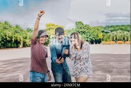 Trois amis joyeux regardant le téléphone portable faisant geste gagnant à l'extérieur. Amis adolescents excités regardant l'écran de téléphone portable dans la rue Banque D'Images