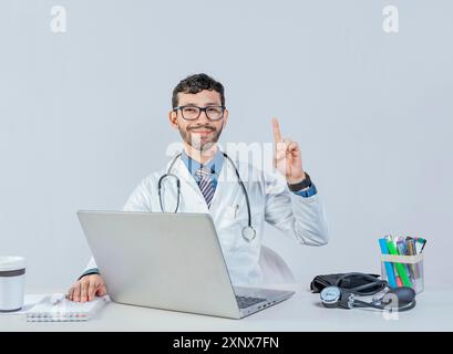 Médecin souriant avec ordinateur portable avec doigt vers le haut dans le geste de l'idée. Médecin assis avec un ordinateur portable pointant vers le haut isolé. Jeune médecin assis avec un ordinateur portable avec un Banque D'Images