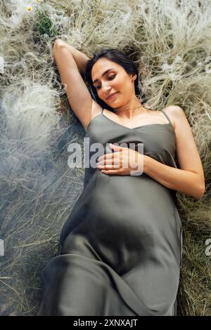 Jeune femme enceinte en robe de satin vert repose sur l'herbe en été. Charmante future mère tient doucement son ventre dans la prairie. Femme paisible attendant Banque D'Images