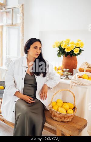 Jeune femme sourit et s'assied à table avec des citrons. Pichet en argile avec roses jaunes, plante d'intérieur, ustensiles de cuisine sur nappe. Calme brune fille i Banque D'Images