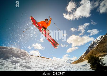 Beau jeune homme caucasien dans une veste orange et un short noir sautant d'un tremplin sur des skis rouges sur fond de montagnes, ciel Banque D'Images