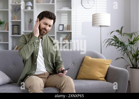 Homme assis sur le canapé éprouvant des douleurs à l'oreille tout en tenant le smartphone en main. Il a l'air mal à l'aise et souffre. Décor moderne visible en arrière-plan. Banque D'Images