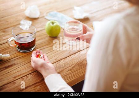 Angle élevé de la femelle anonyme avec des pilules et une tasse d'eau en mélange à table avec du thé et de la pomme pendant la maladie Banque D'Images