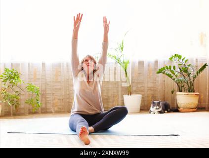 Femme adulte dans des vêtements décontractés, levant les bras et étirant le corps tout en faisant du yoga sur le sol à la maison Banque D'Images