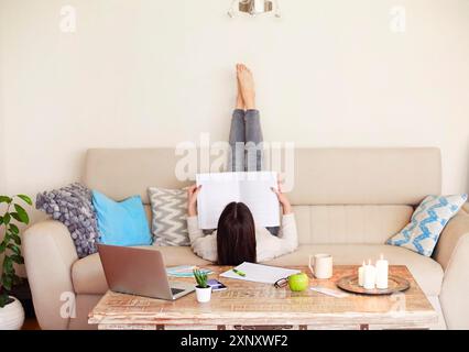 Vue arrière de jeune femme pieds nus méconnaissable dans une tenue décontractée couchée sur un canapé avec les jambes vers le haut et la lecture de la littérature professionnelle tout en travaillant Banque D'Images