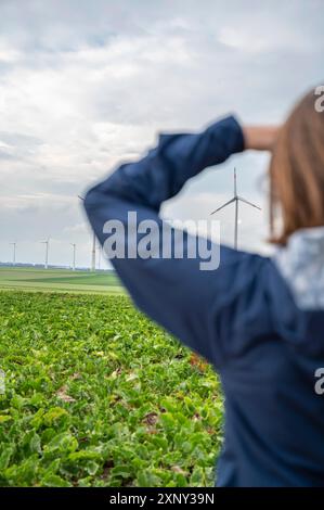 Femme aux cheveux bruns et blouson bleu regarde un parc éolien à distance, parc éolien avec éoliennes dans la zone agricole, vue arrière Banque D'Images