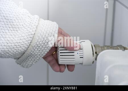 Main d'une femme dans des vêtements chauds réduit la chaleur au thermostat sur le radiateur en raison de la crise énergétique avec la hausse des prix, l'espace de copie, sélectionné Banque D'Images