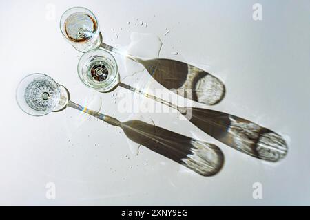 Trois verres différents remplis d'eau projettent de longues ombres sur un fond gris et de petites flaques dans lesquelles la lumière est réfractée Banque D'Images