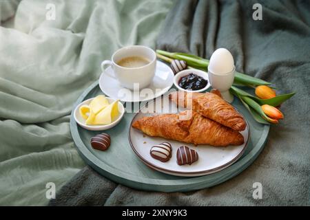 Petit déjeuner servi avec amour au lit, café, croissant, confiture, oeuf, quelques coeurs en chocolat et tulipes sur un plateau sur le linge de lit vert gris à un romantique Banque D'Images