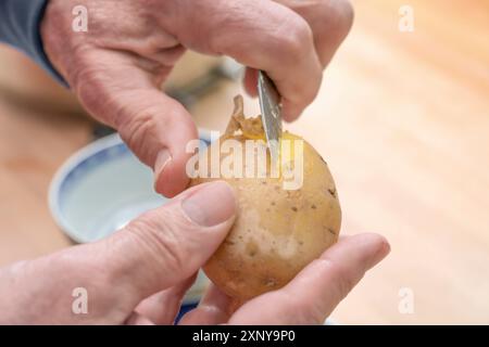 Les mains d'un homme âgé épluchant une pomme de terre cuite avec un couteau de cuisine, préparant des aliments, un espace de copie, un foyer sélectionné, une profondeur de champ étroite Banque D'Images