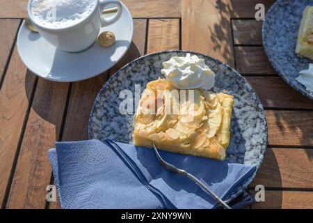 Gâteau aux pommes frais recouvert d'amandes, servi avec de la crème fouettée sur une assiette bleue et une tasse de café sur une table de jardin en bois, le jour du soleil Banque D'Images