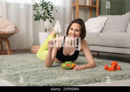 Belle jeune femme heureuse avec bol de salade fraîche et haltères couchés à la maison. Concept de perte de poids Banque D'Images