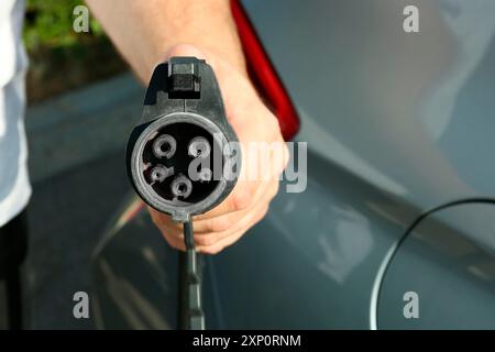 Homme tenant le câble d'alimentation près de la voiture à la station de charge de véhicule électrique, gros plan Banque D'Images