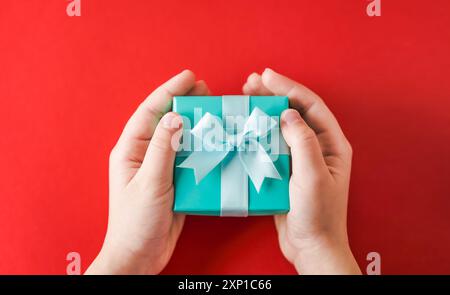Mains d'enfant tenant un cadeau turquoise ou une boîte-cadeau sur un fond rouge, vue de dessus, pose plate. Banque D'Images
