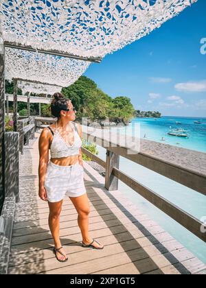 Une femme se tient sur une promenade en bois surplombant l'océan. Elle porte un débardeur blanc et un short Banque D'Images