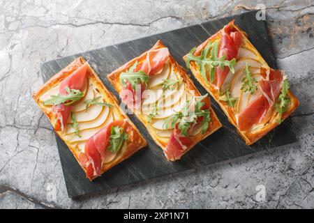 Grignoter des sandwichs feuilletés ouverts avec poire, jamon et roquette en gros plan sur une planche de marbre sur la table. Vue horizontale de dessus Banque D'Images