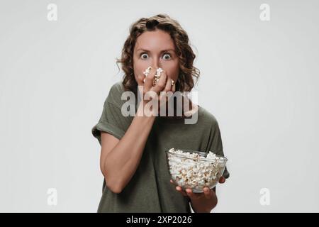 Jeune femme avec les cheveux bouclés mangeant du pop-corn, regarder un film d'horreur avec une grande surprise, regarder la télévision, isolé sur fond blanc. Copier l'espace Banque D'Images