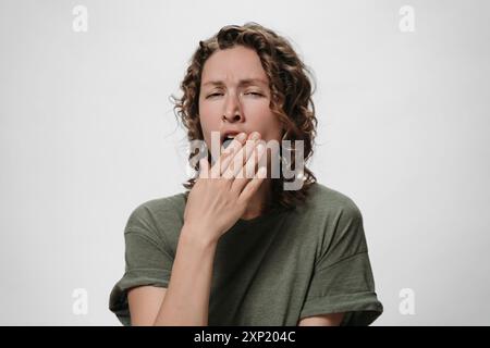 Femme endormie bouclée fatiguée bâille et couvre la bouche avec la paume, l'air ennuyé, se tient sur fond blanc avec un espace vide pour votre contenu promotionnel Banque D'Images