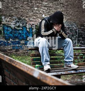 Jeune homme en détresse, assis sur un banc dans une cour de Berlin-Kreuzberg couverte de graffitis. L’image représente la vie urbaine et les luttes de santé mentale. Banque D'Images