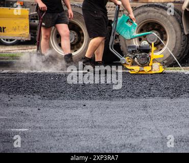 Vue rapprochée des travailleurs utilisant un compacteur pour lisser et niveler l'asphalte fraîchement posé sur un chantier de construction de voie ferrée. Banque D'Images