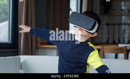 Un jeune garçon engagé dans une expérience de réalité virtuelle à la maison. Il porte des lunettes VR et a les bras tendus, semblant naviguer ou interagir avec Banque D'Images