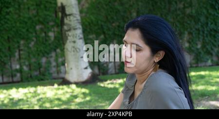 Une femme aux cheveux longs est assise dans un parc. Elle porte une chemise verte et a la tête inclinée Banque D'Images